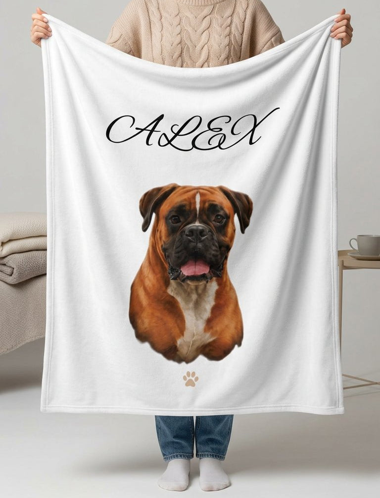A woman holds up a white personalized blanket featuring a printed boxer dog portrait and the name 'ALEX' in black script. The setting is a clean, modern bedroom with a bed and side table in the background.